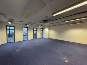 Empty office space on De Ruijterkade with blue carpet, white walls, and large windows facing the street.